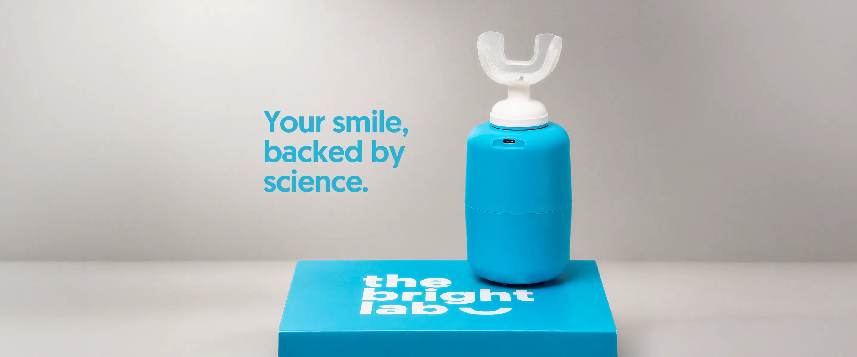 The Bright Lab applicator on its charging base, standing on a branded box: your smile, backed by science